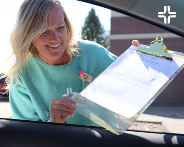 Smiling healthcare worker in a teal sweatshirt leaning toward a car window while holding up a clipboard with paperwork for a driver.